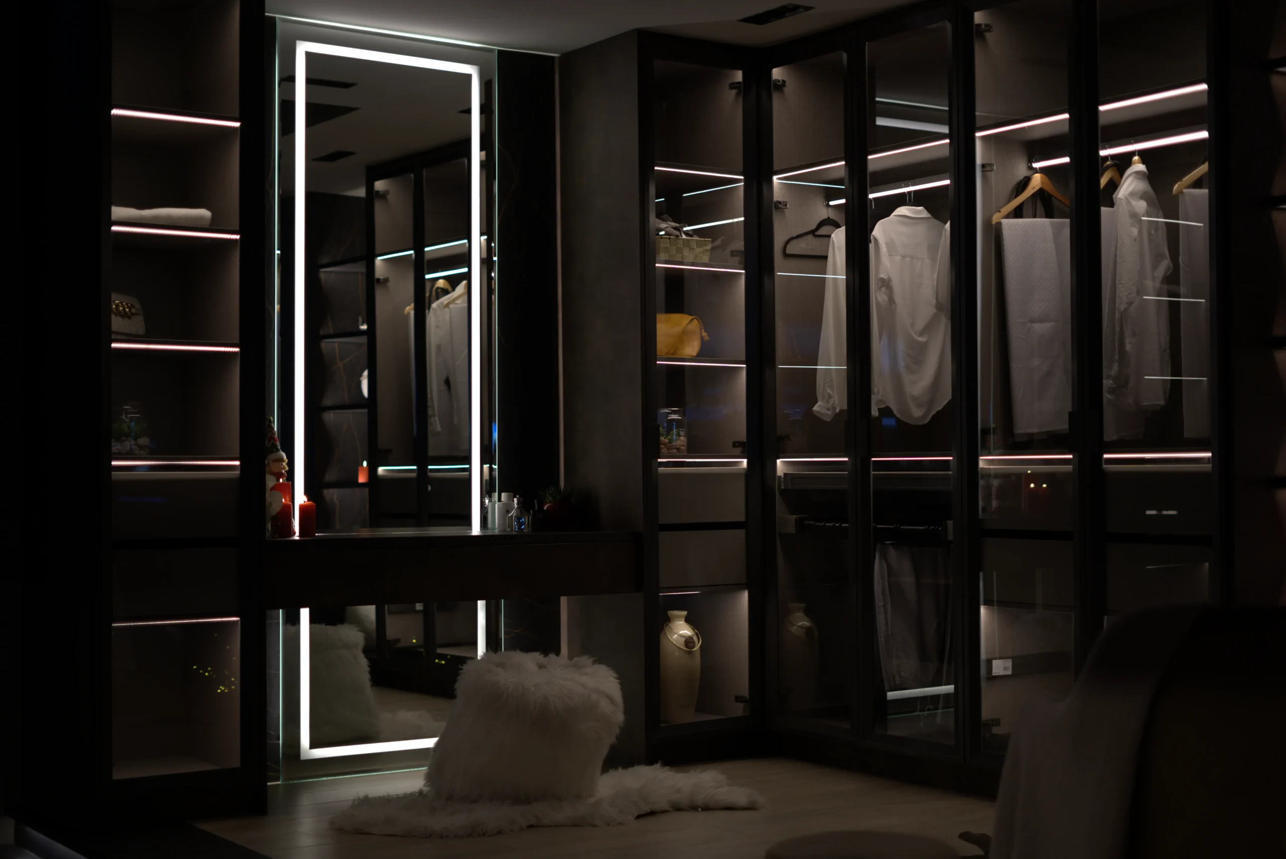 Luxury walk-in closet with glass wardrobe doors, LED strip lighting, white shirts on hangers, a vanity with a backlit mirror, and a fluffy white stool on a rug.